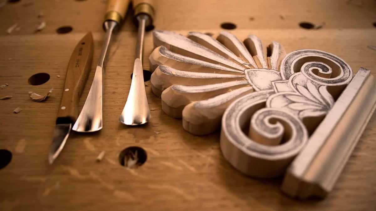Close-up of carving chisels resting beside a beautifully detailed wood piece. The workbench is marked with shavings, showing the hands-on effort in wood carving classes and lessons. These tools are essential for anyone looking to hone their carving skills and create stunning designs.