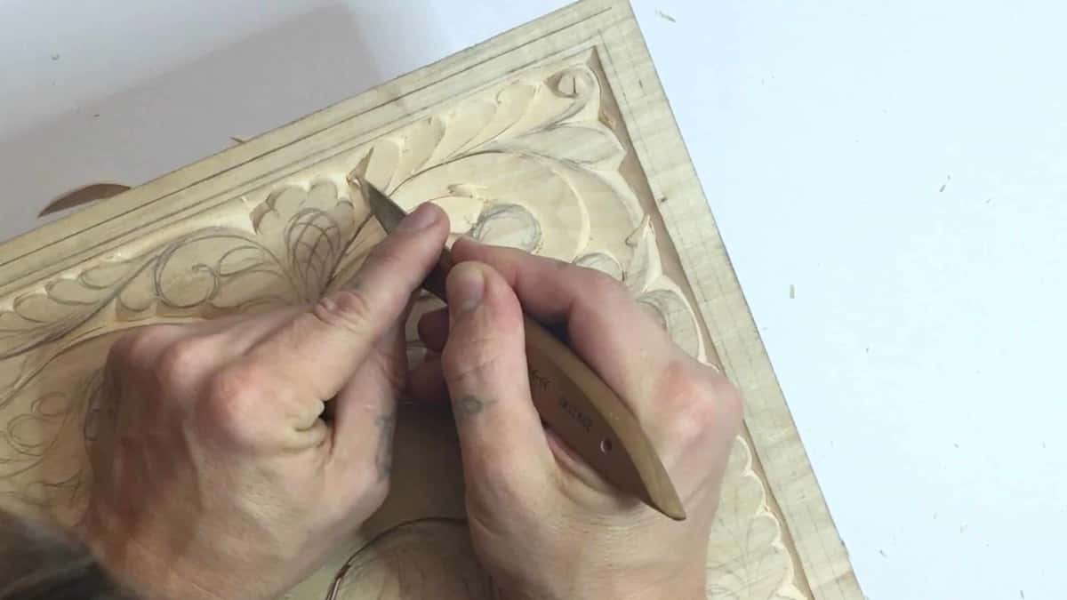 Both hands steer a wood handled carving knife into a penciled leaf on a pale basswood panel.