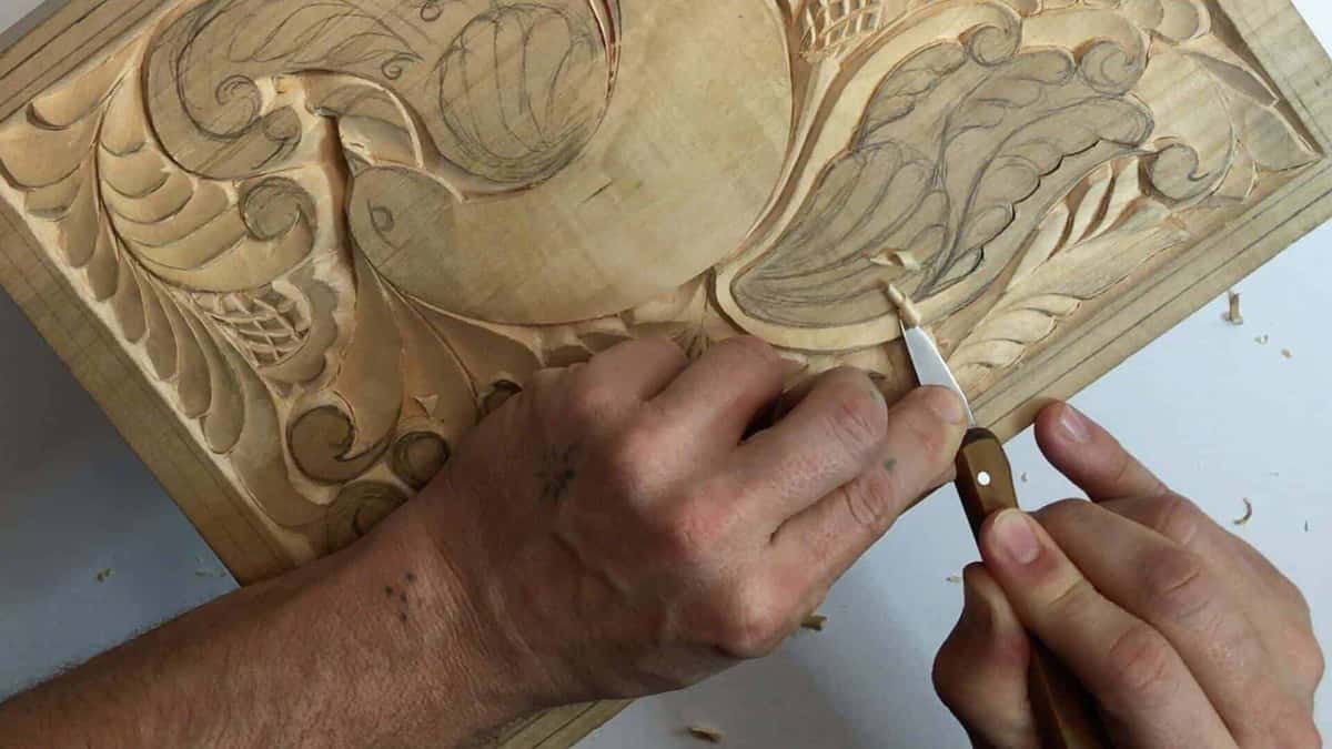 Fine shaving curling off a pale basswood panel under a small skew, pencil work still showing