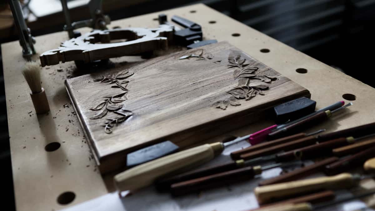 A walnut board with a carved leaf border rests on the bench beside a row of gouges and a dust brush