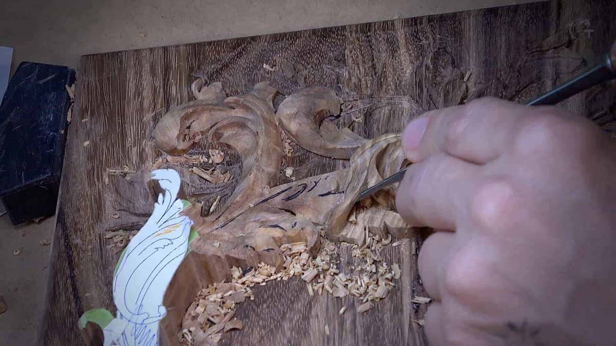 Overhead of a walnut panel, a gouge shaping the acanthus scroll while fresh chips pile up beside it