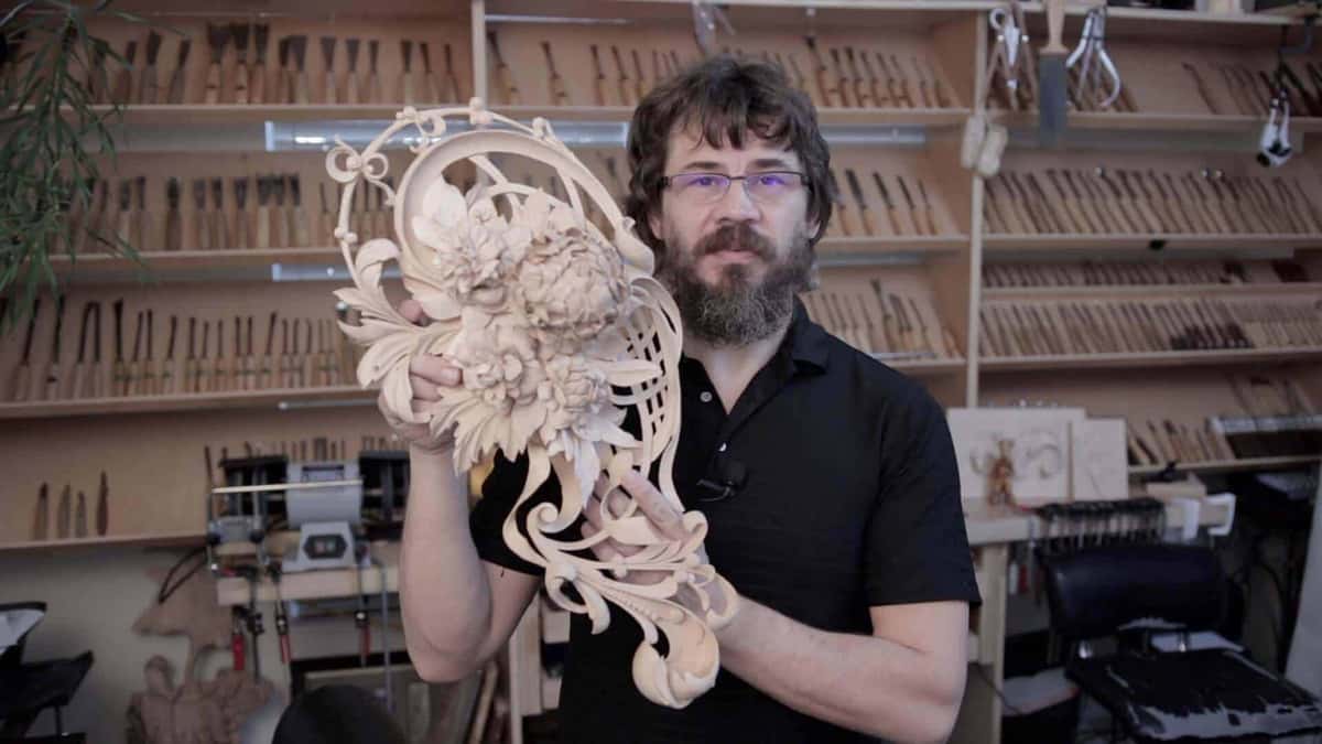 A man holds a big pierced flower carving; his shop walls are racked with gouges behind him.