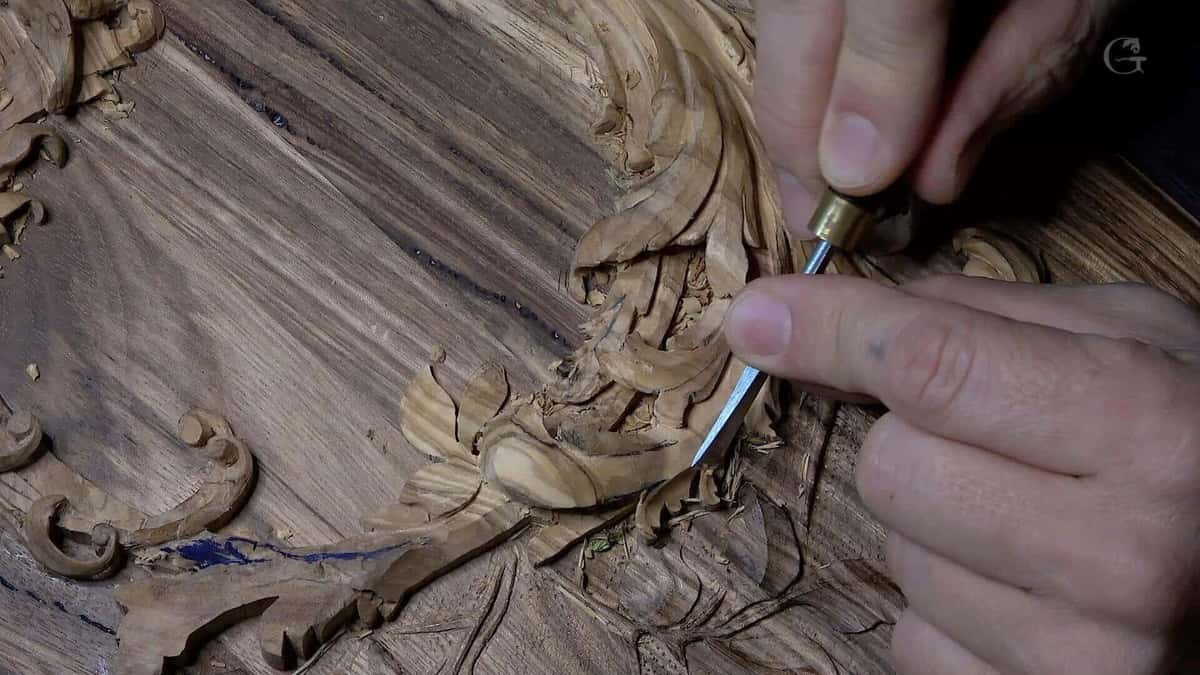 Two hands steer a small brass ferruled gouge through tight figured grain on the lily box