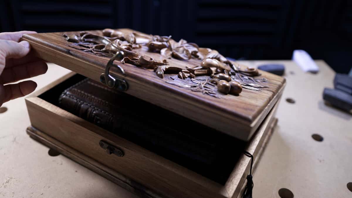 A hand lifts the carved lid off a small box with a leather bound book inside