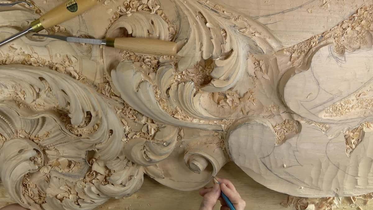 Overhead view of a large rococo panel part carved, two gouges lying across it, a blue pencil in hand.