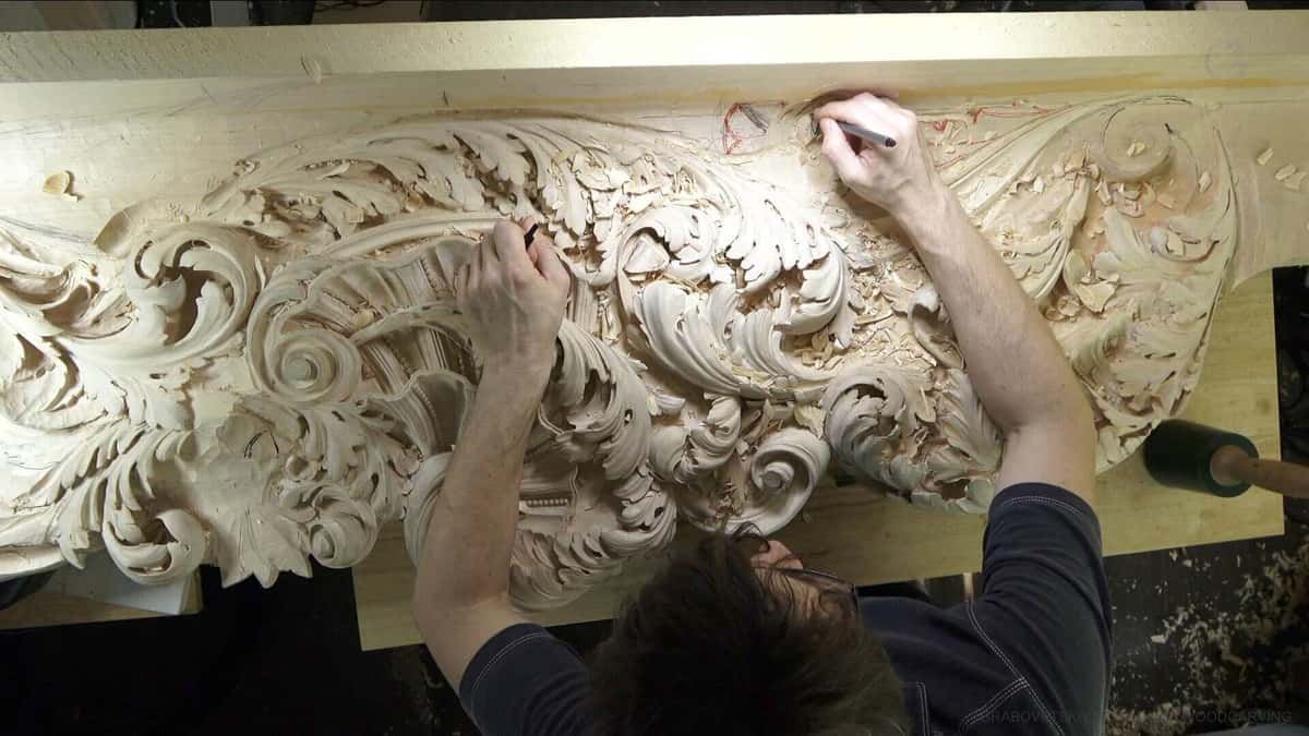 Both hands drawing pencil lines back onto a half-carved acanthus frieze, red and graphite marks on the wood
