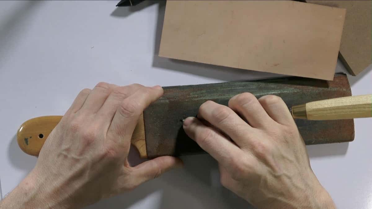 Hands holding a flat strop board loaded with green polishing compound, a wood handled gouge resting on it ready for honing