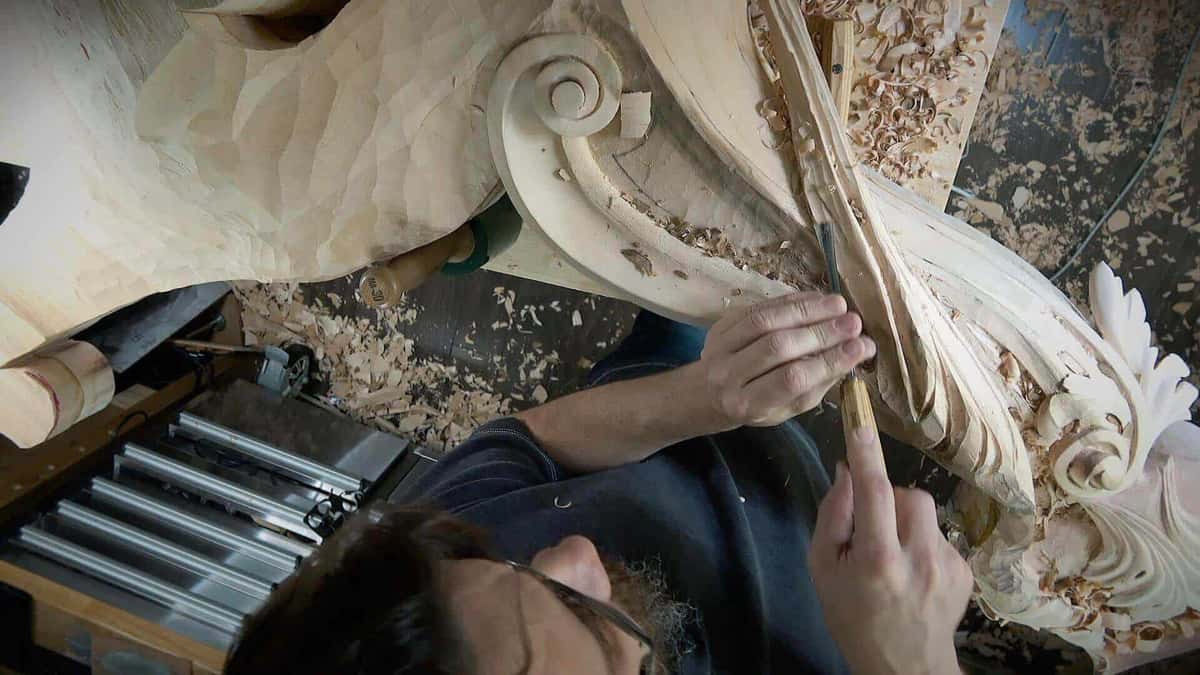 Seen from above, both hands guide a gouge along a big pale scroll while shavings pile on the floor.
