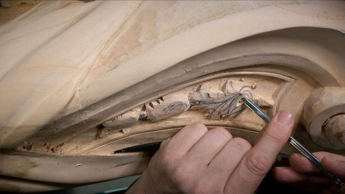 The image shows a close-up of a hand holding a carving tool, working on intricate wood carvings. The wood surface features detailed, flowing designs that appear to be in the process of being refined or completed. The focus is on the craftsmanship involved in wood carving.