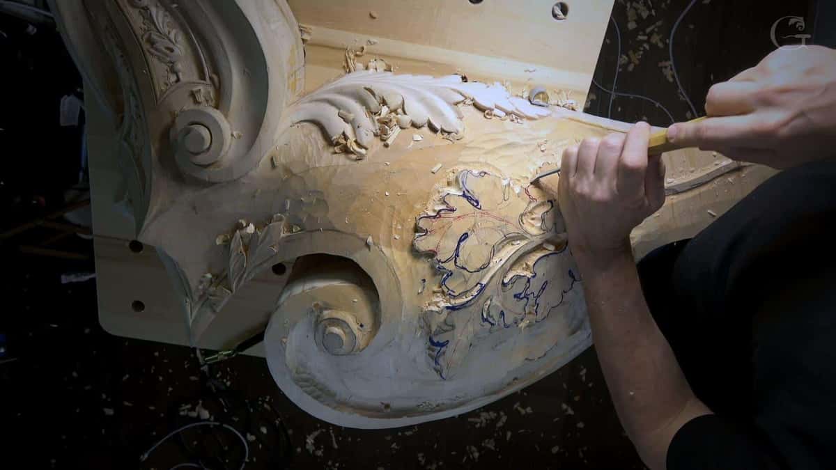 Two hands carving a large architectural scroll in pale wood.