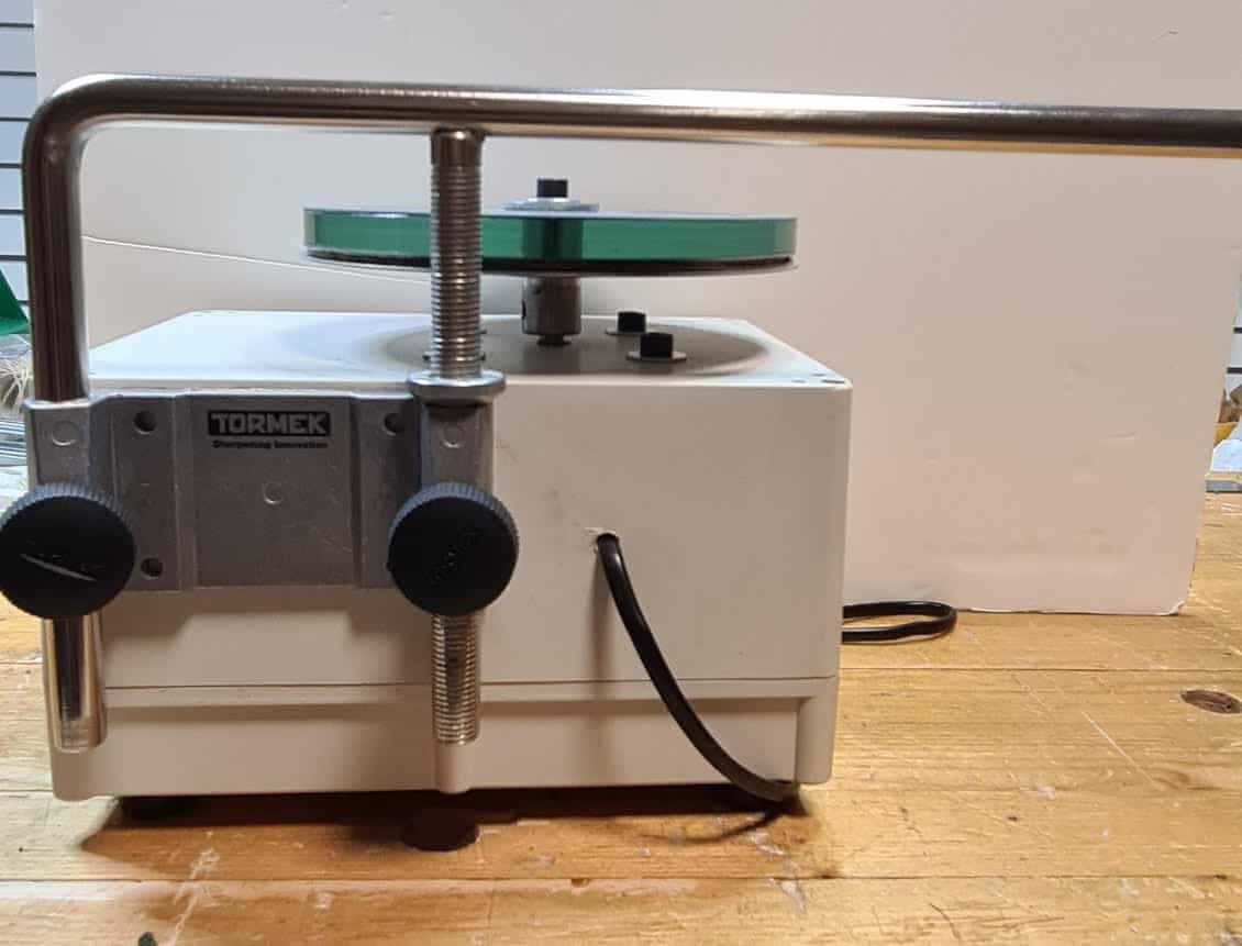 White cased sharpening machine sits on a dusty bench, its chrome jig bar arching over the green rimmed honing wheel