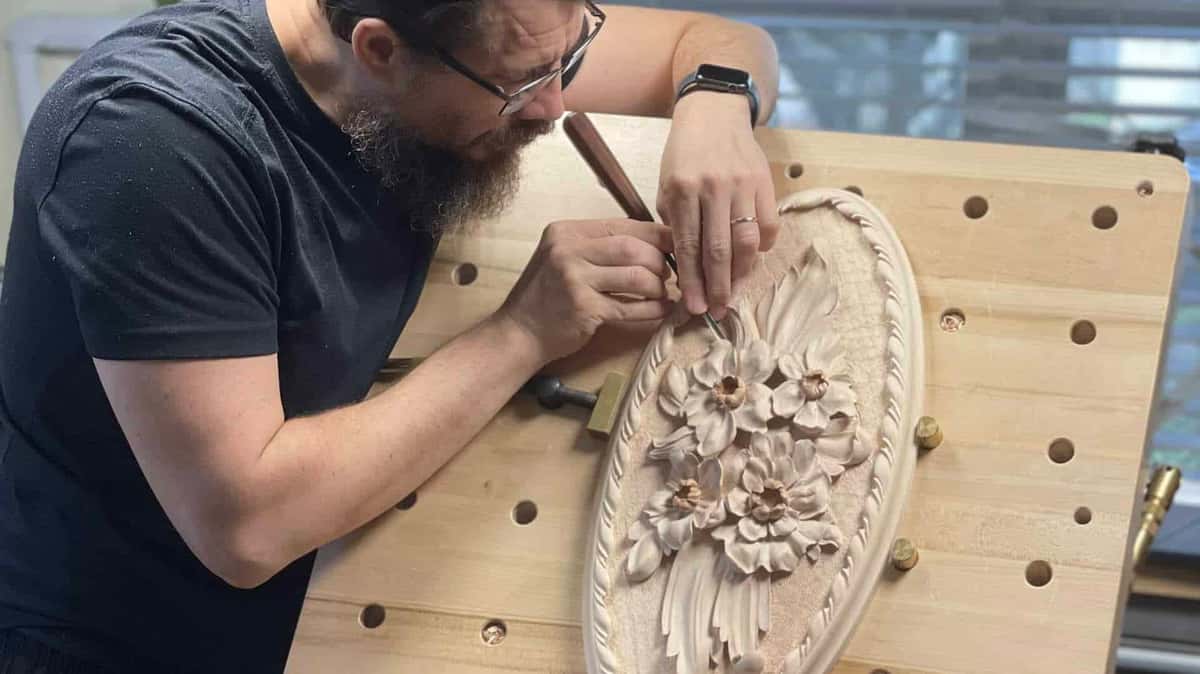 Master carver bent over an oval floral rosette held by bench dogs and a brass screw clamp, small tool in both hands