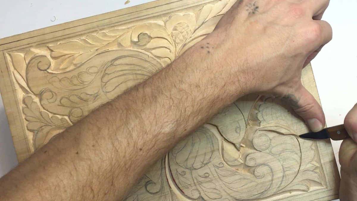 Bare forearm laid across a carved acanthus board while a knife works the drawn area beyond it