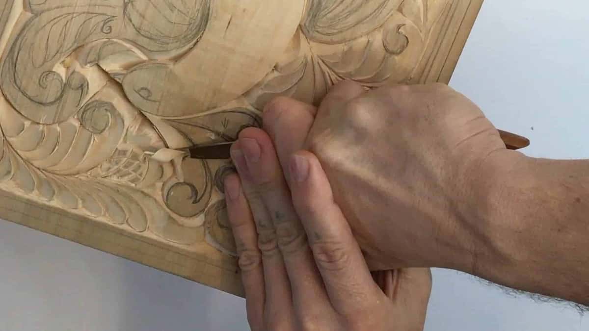 Palm and thumb pushing a small gouge along the grain of a pale relief carving on the bench