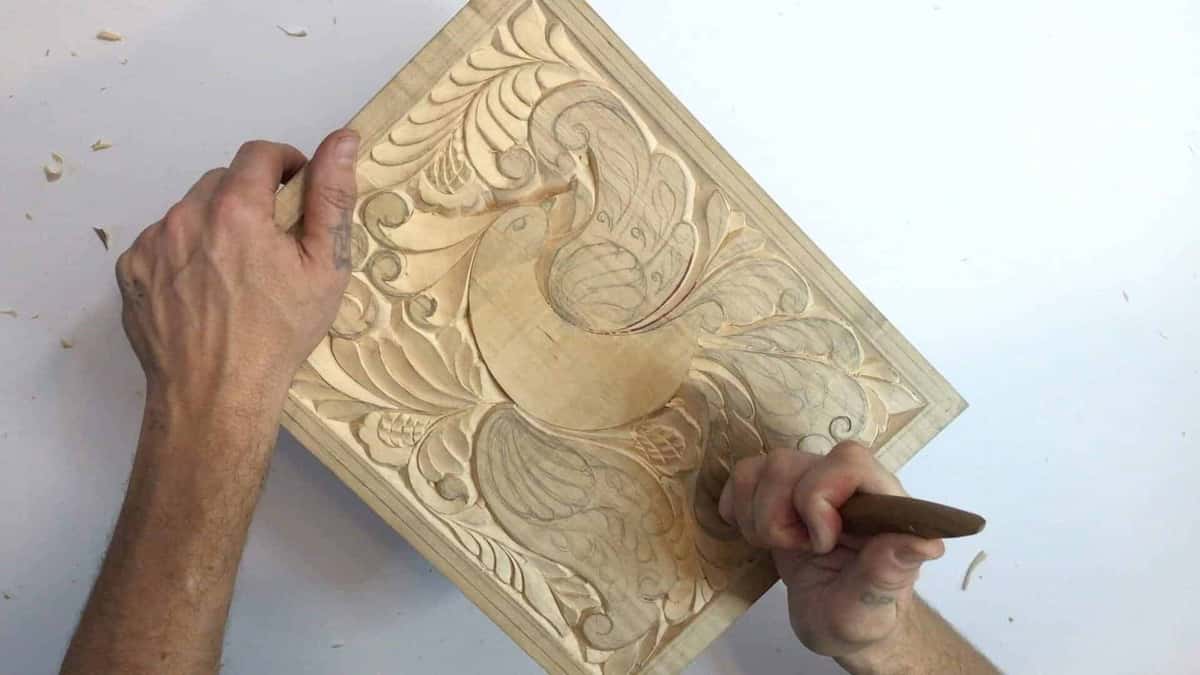 Working hand at the lower right edge of a carved bird panel, the blade hidden inside the fist