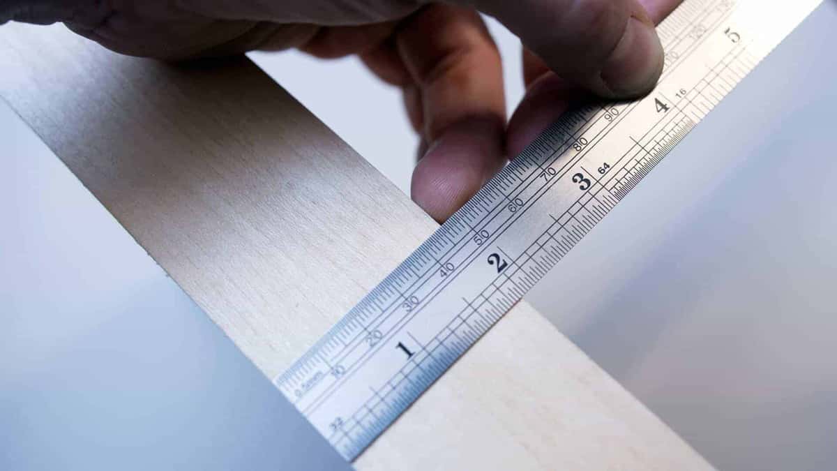 Fingers hold a marked steel rule across a pale wood strip, reading the width straight off the printed scale
