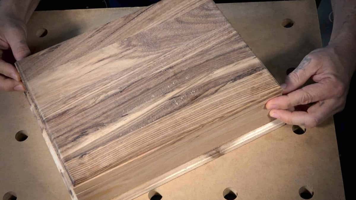 Two hands flattening a striped board of olive and acacia on a dog holed bench
