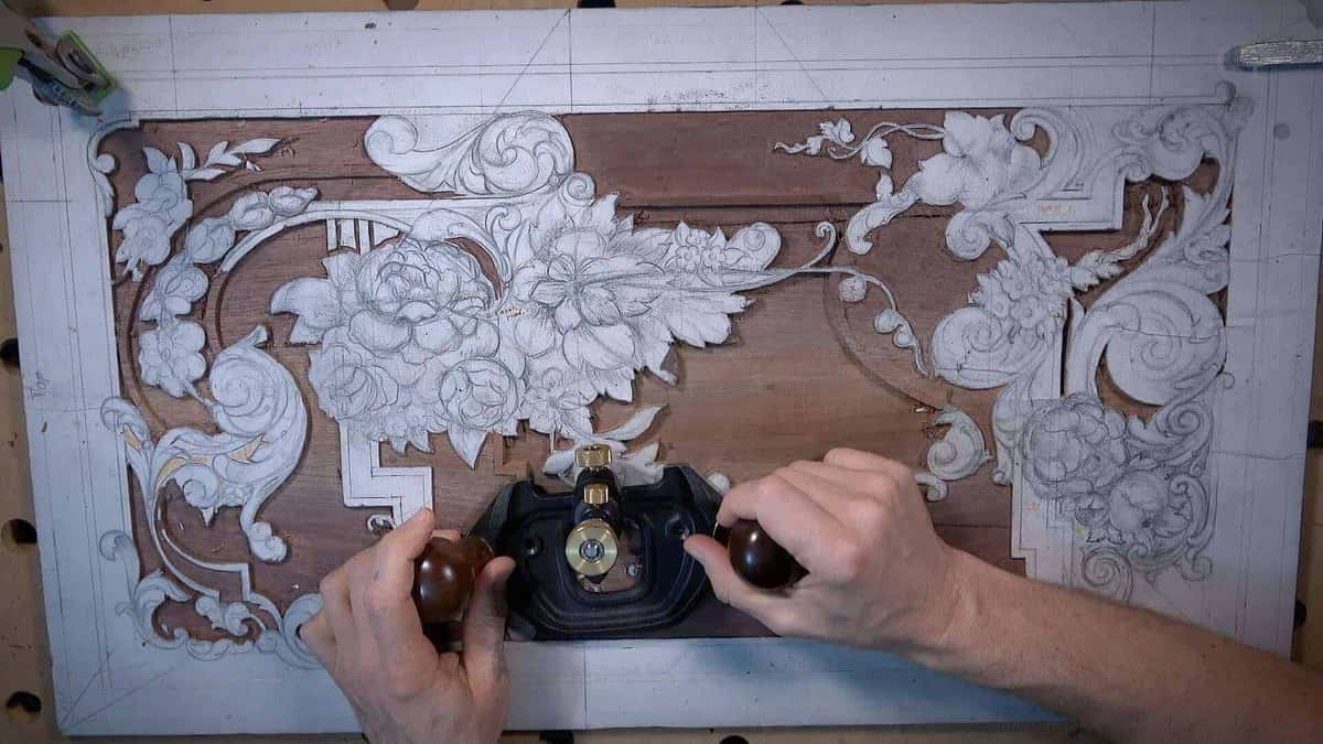 Both hands running a router plane across the sunken ground of a floral furniture panel