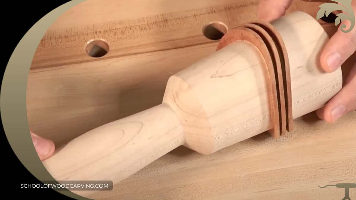 Two hands turning a pale mallet head banded with three copper rings on the bench