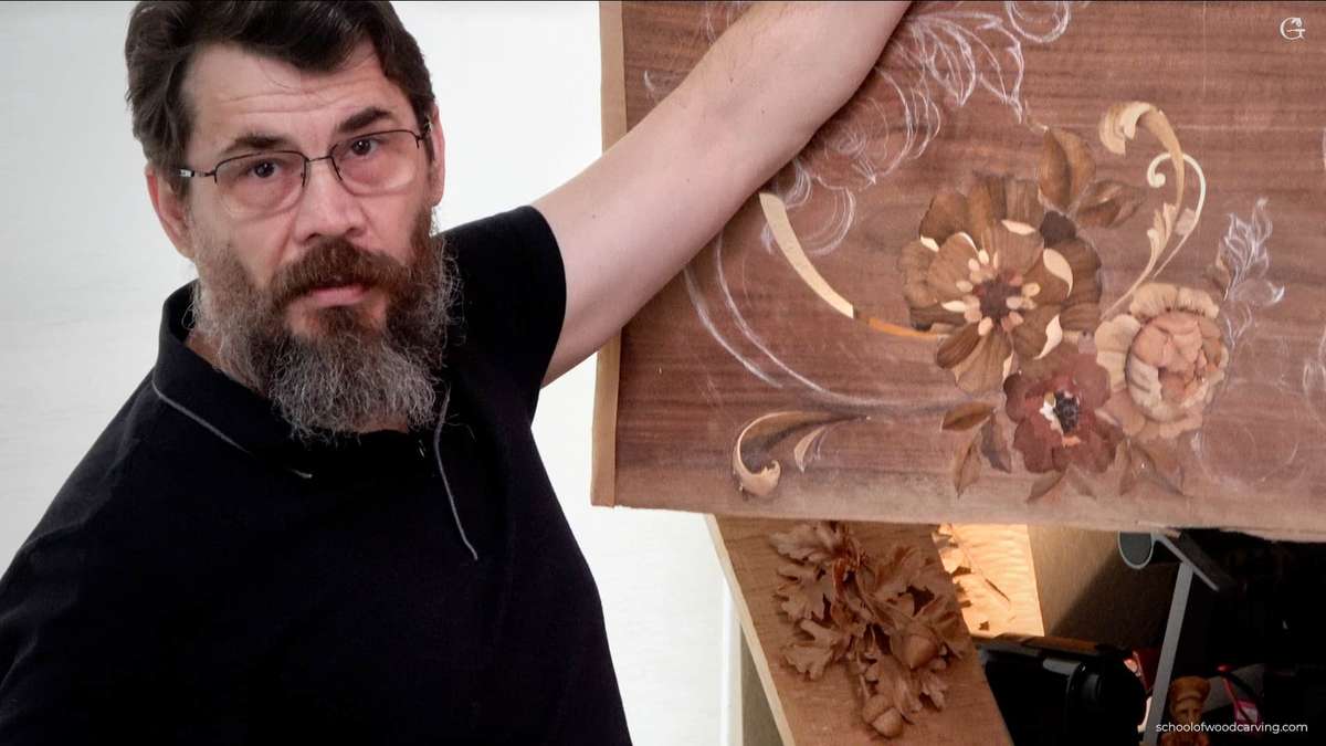 Man with glasses and a beard holding a wooden panel featuring intricate floral carvings and designs.