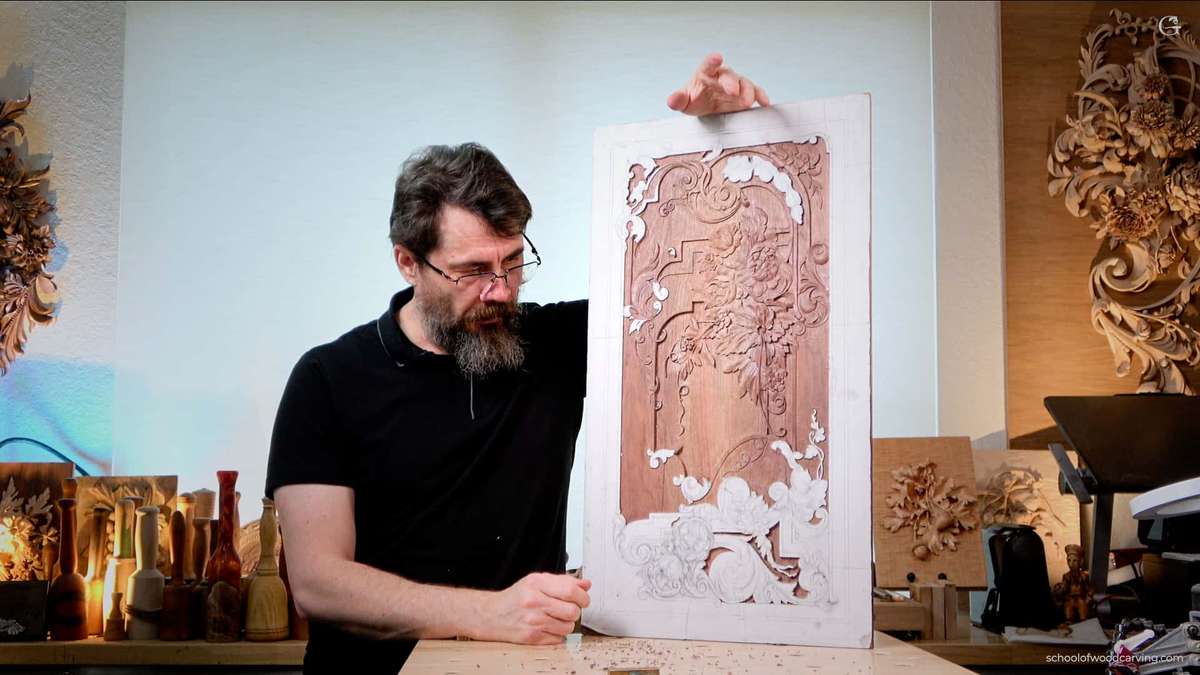 A man with a beard is carving a furniture panel.