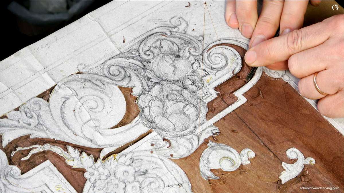 A craftsperson's hands carve a detailed floral design into a wooden panel using precision tools, with a sketch nearby for reference.