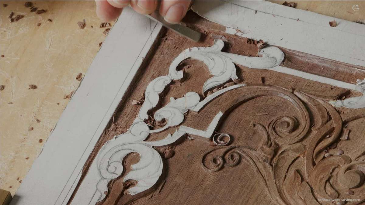 WOODCARVING LESSONS: A hand holds a carving tool and cuts beautiful, swirling shapes into brown wood. The pattern comes from a paper outline, which is still on the wood to help guide the work. You can see small pieces of wood, called shavings, around the area. Only Woodcarver Grabovetskiy is in the picture. Learn more about this step-by-step woodcarving lesson or course with Woodcarver Grabovetskiy. This simple tutorial shows how to use patterns and tools for beginner and advanced carving projects. - Wood Carving tutorials