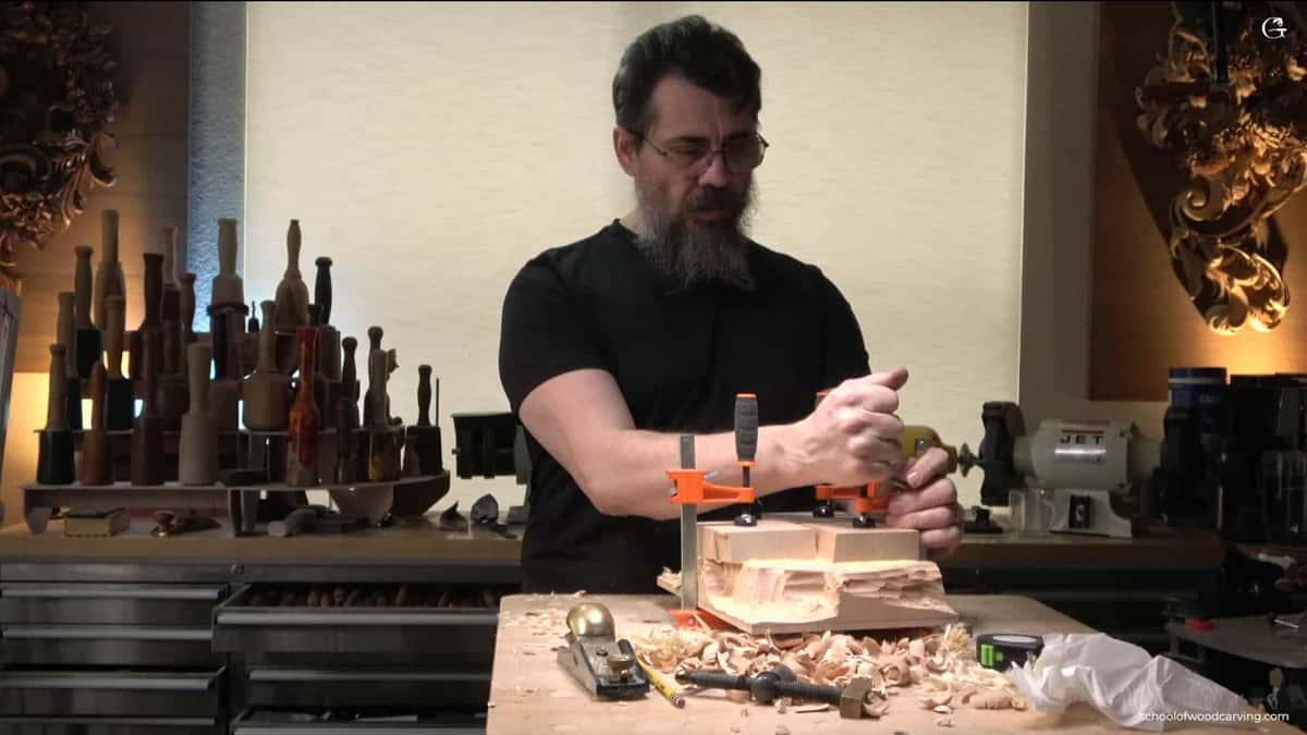 Two clamps press down on the thick square blocks left at the top of a roughed panel, well clear of the carving