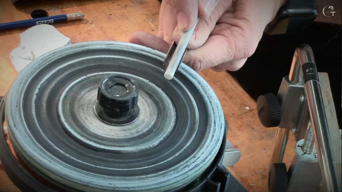 A gouge held to a honing wheel on a scarred orange workbench, a marking pencil and a jig beside it.