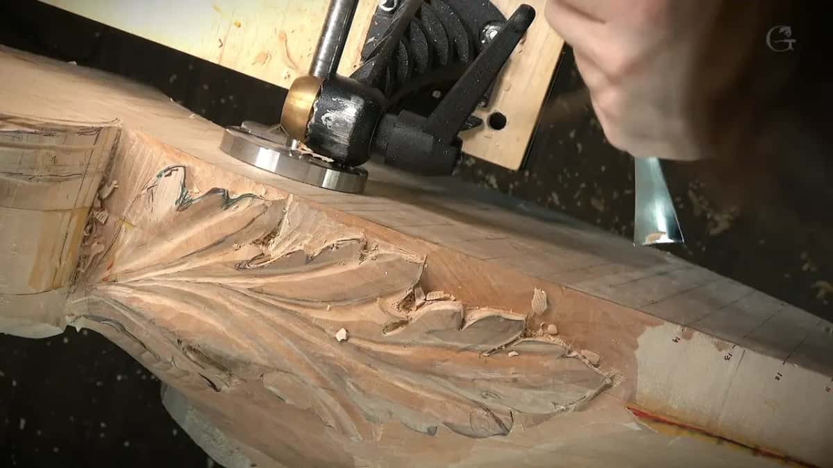 Clamped upright in a bench vise, a carved acanthus leaf panel is dusted with wood shavings, a wide chisel resting nearby.