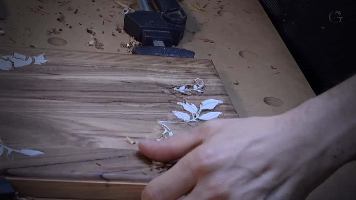 Straight wood grain runs across a board on the workbench, a white paper leaf pattern taped on and a hand nearby.