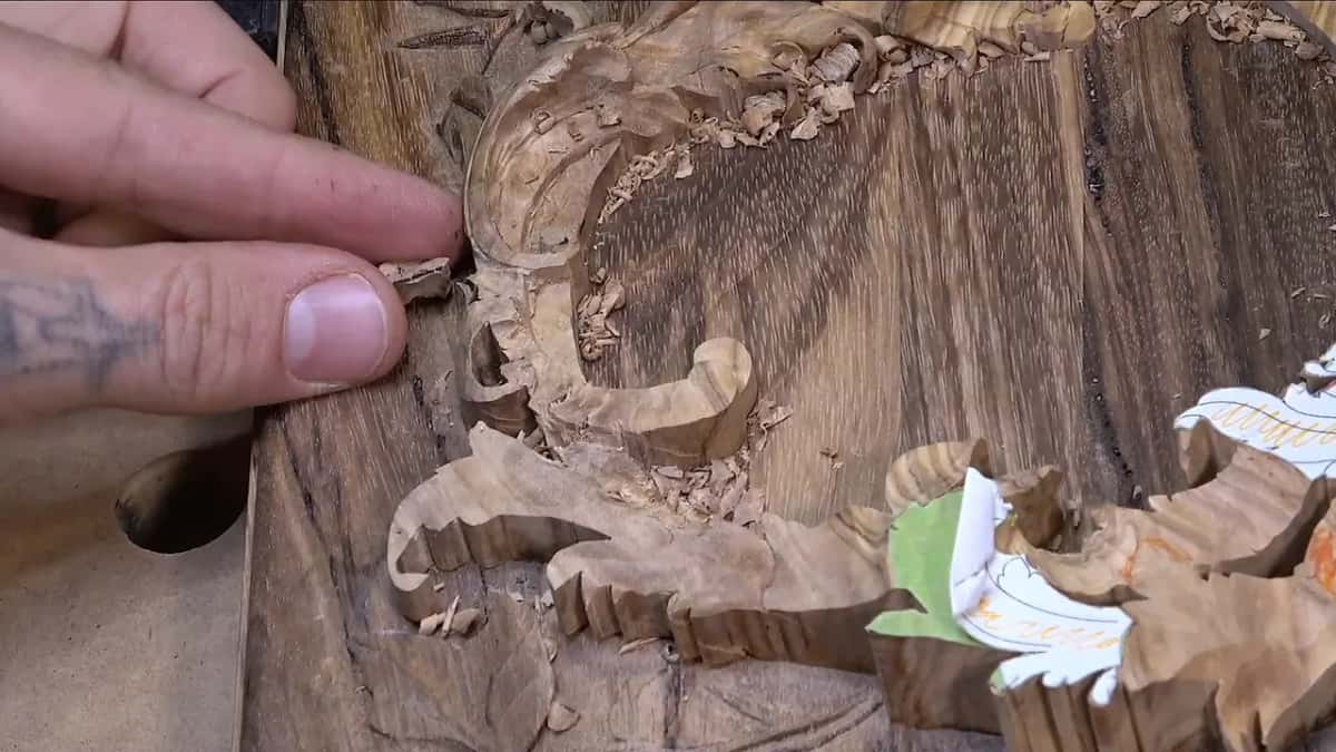 Fingers press a small broken wood piece back against a carved scroll, with shavings and a paper pattern nearby.