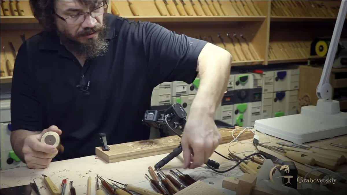 In his workshop, a bearded carver holds a round tool in one hand while reaching for others on the bench.
