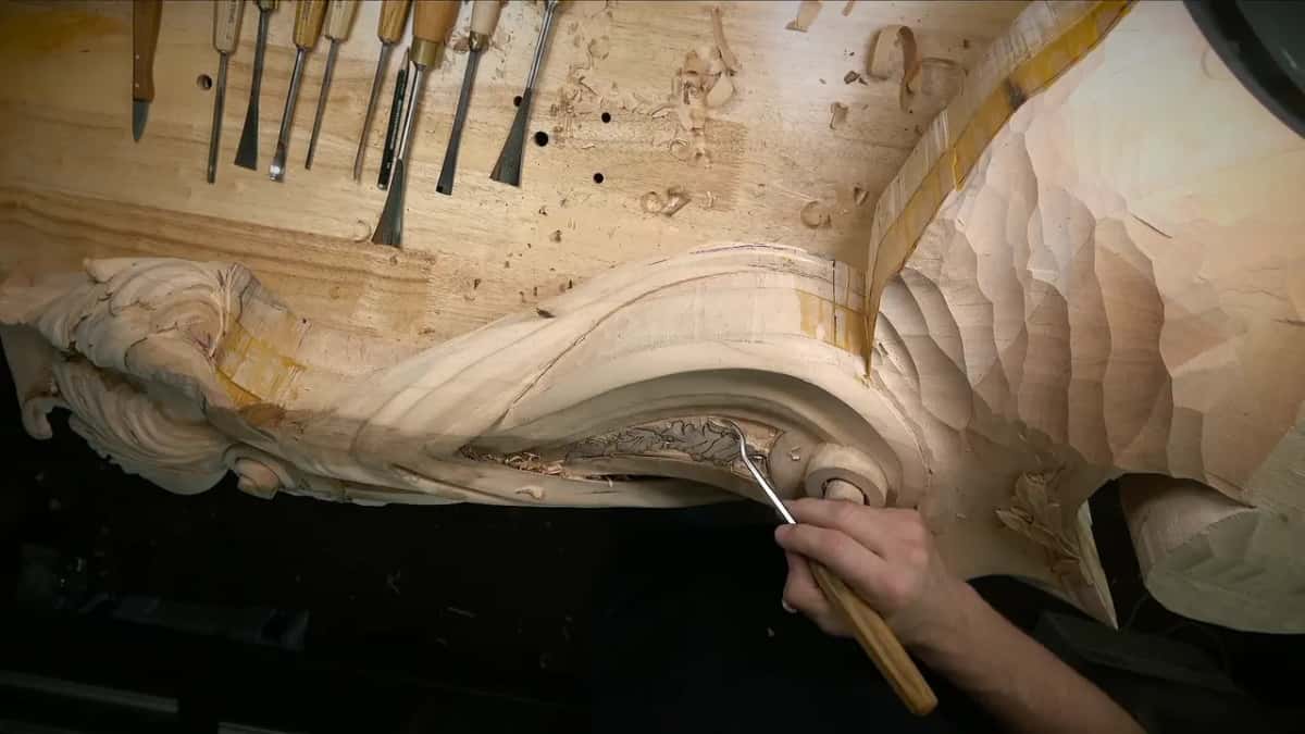 Under a gouge, a large scrolling wood bracket takes shape, a strip of tape marking a line, tools lined up above.