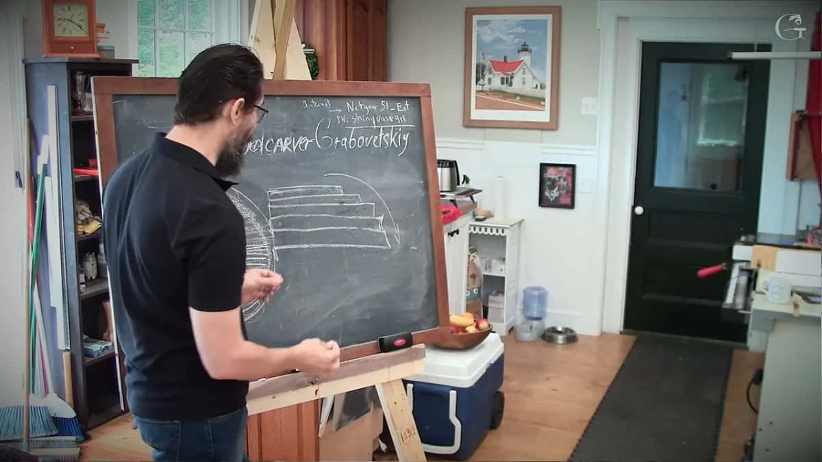 At a blackboard, a bearded man draws a layered, curved diagram in chalk.