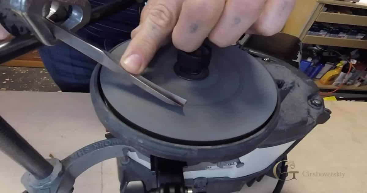 A hand presses a flat chisel onto the dark wheel of a grinder, the guide arm sitting at the left.