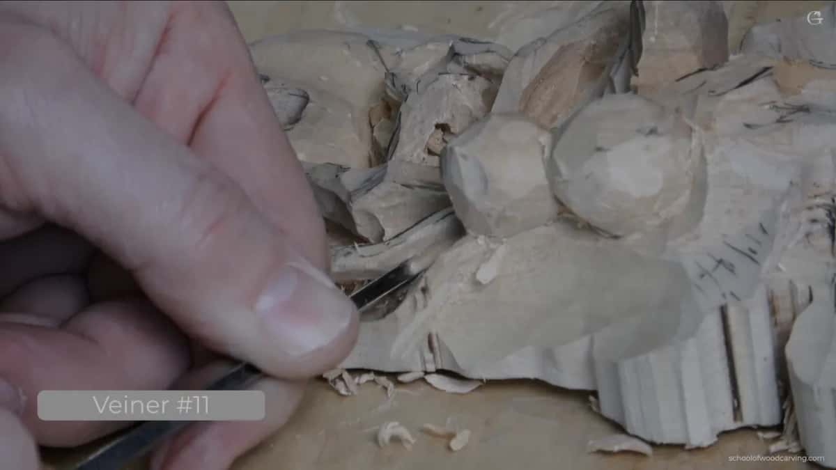 Fingers grip a narrow gouge working into round bead shapes on a roughed wood carving, chips scattered near the blade.