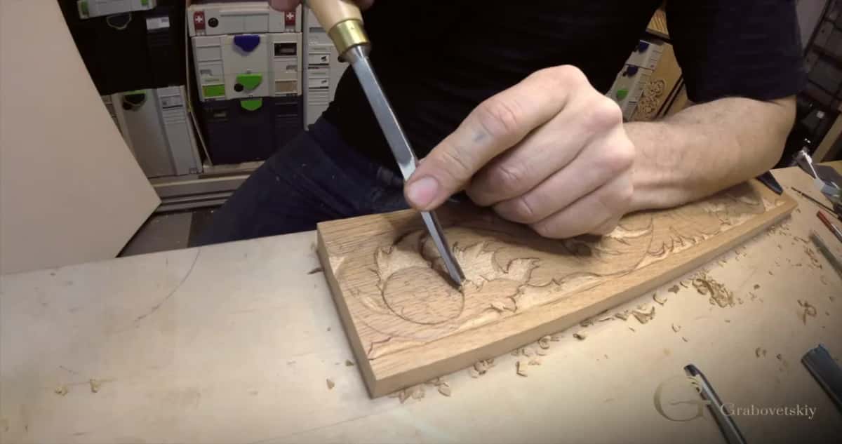 A long chisel stands upright in a tan board where a rosette is half carved, chips scattered on the bench.
