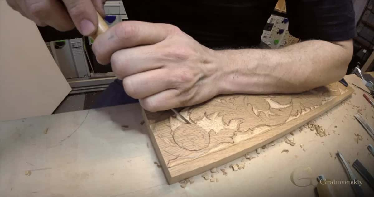 Close in, one hand walks a slim tool along the leaf border of a warm tan board while chips lie beside it.