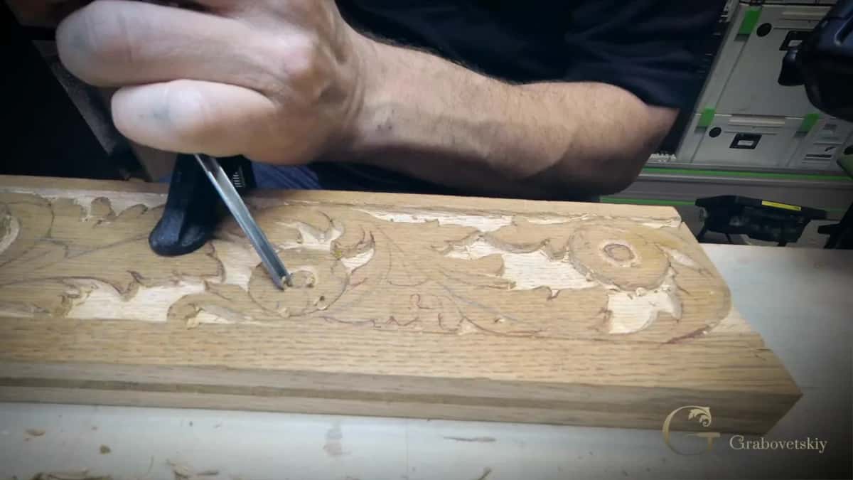 The open grain of this pale board runs straight through the carved leaves, and a thin tool sits in one of them.