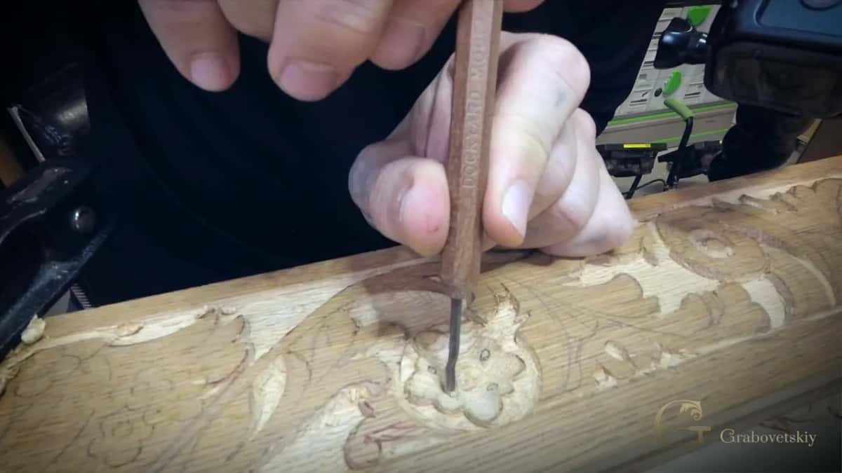 Pressed straight down, a long thin tool sinks into the middle of a carved flower on a pale open grained molding.