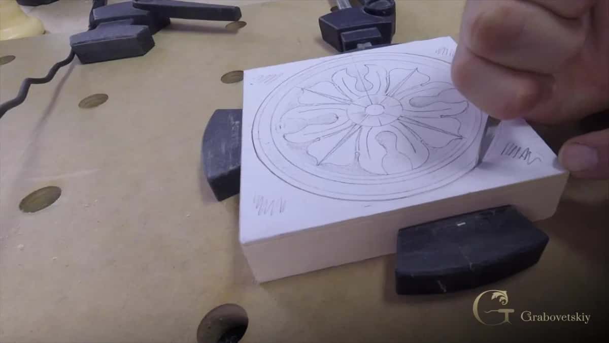 Fingers hold a fine blade at the edge of a creamy block where the rosette pattern is still only pencil.