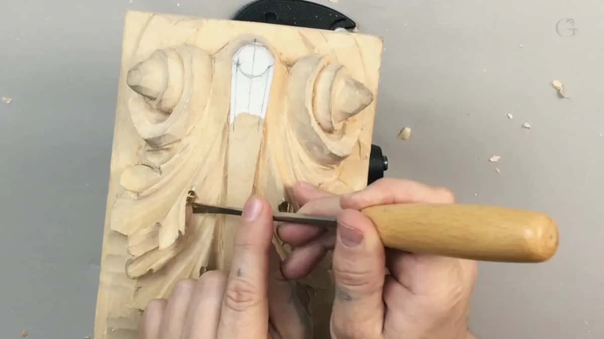 Two hands push a long gouge across a pale panel and the wood peels up in a ribbon ahead of the blade.