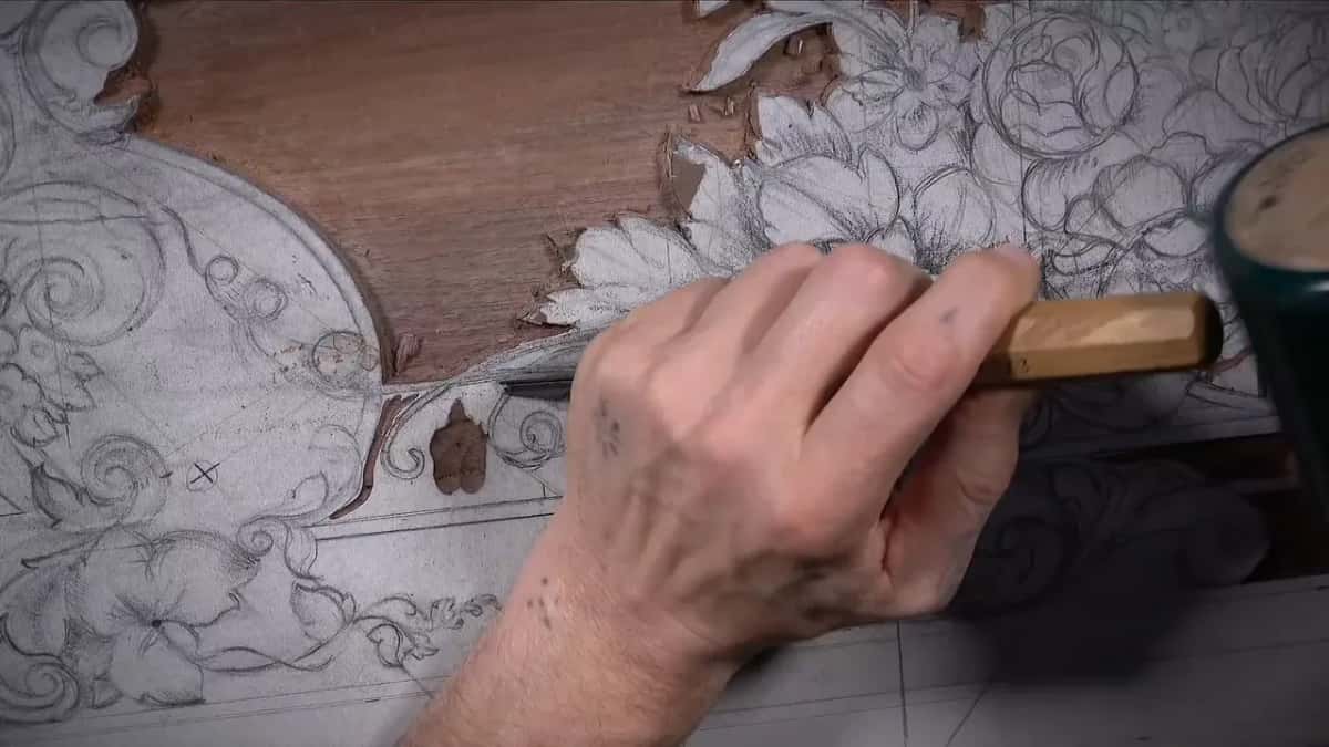 Beside a pencil drawing of flowers, a hand grips a chisel by its light handle over the white dusted panel.