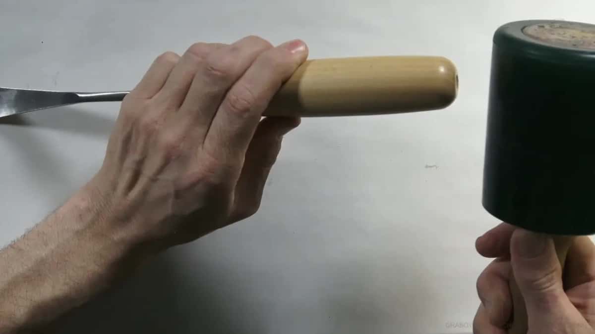 Held ready in one hand, a chisel waits while the other hand brings a green mallet in to strike it.
