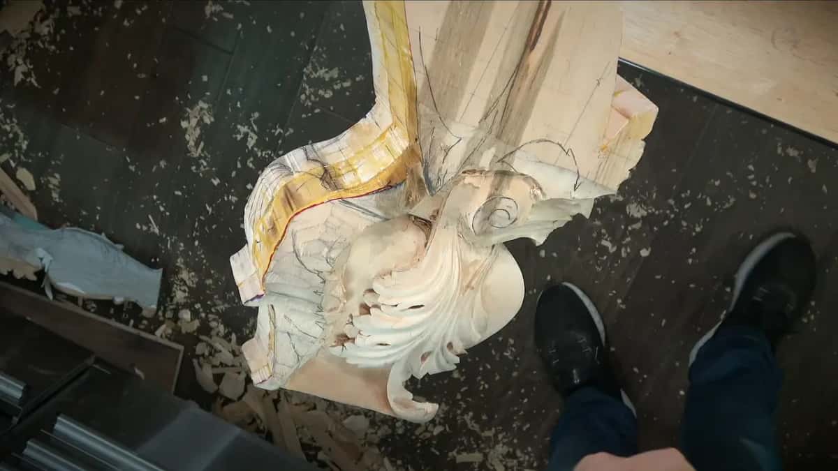 Looking down from the carver's own view: a pale bracket clamped upright, chips over the dark floor and his shoes.