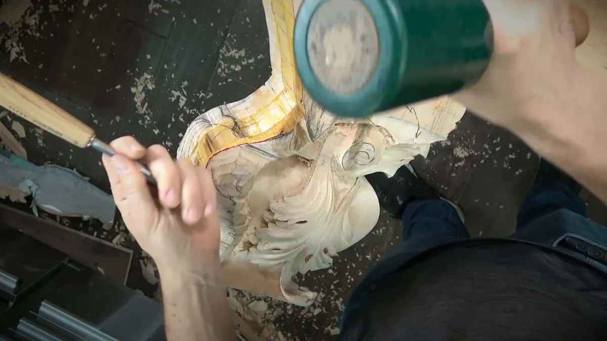 From above, a mallet blurs mid swing toward the gouge held against a pale bracket covered in pencil lines.