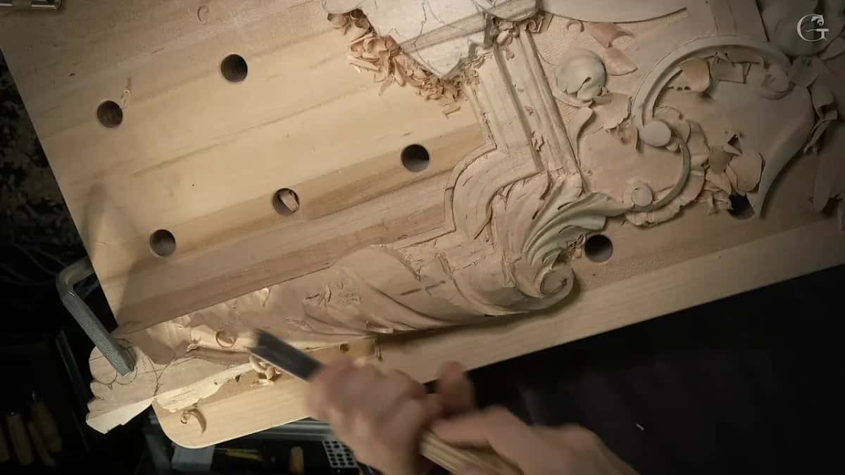 Both hands drive a wide chisel into the lower edge of a panel whose upper ornament is already carved