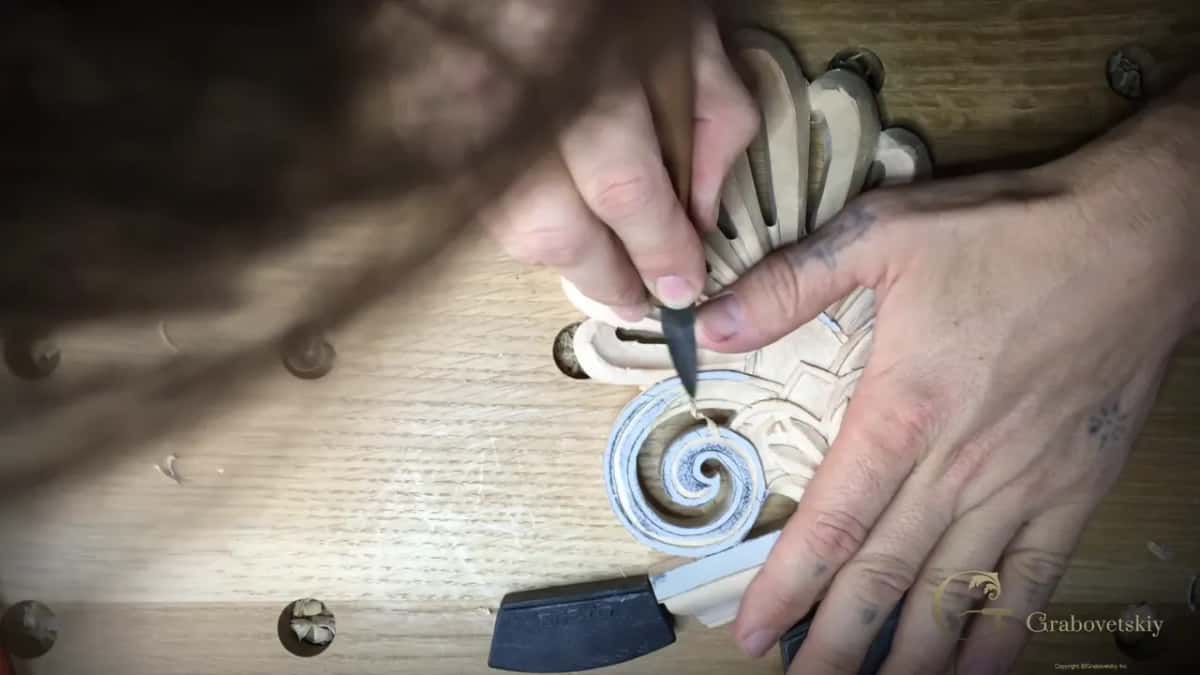 Knife tip working the eye of a chalked spiral volute, both hands steady on the fan-shaped acroterion