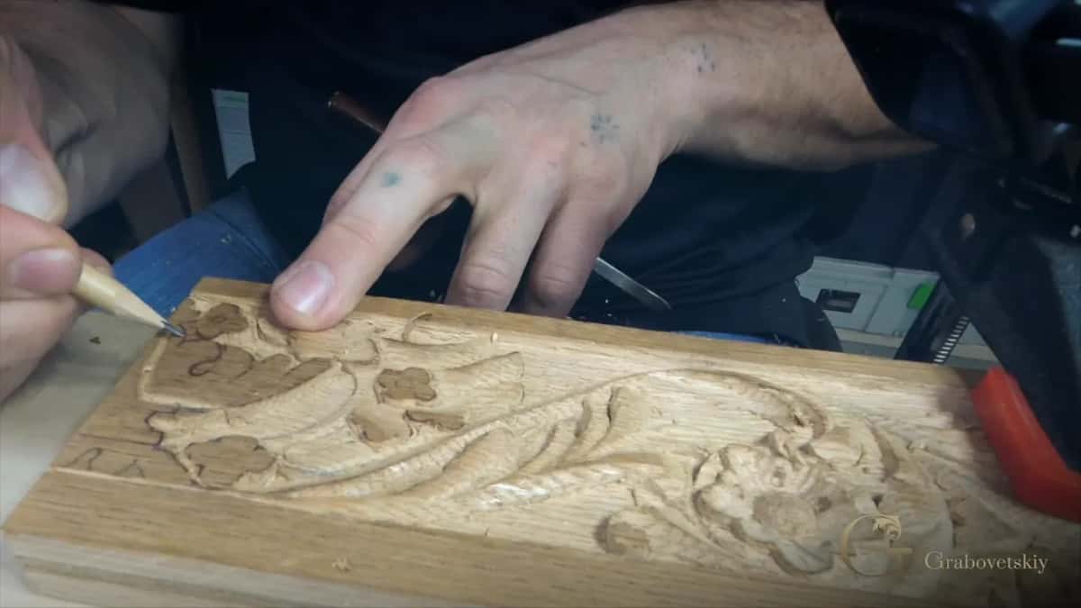Small V-tool outlining a flower on an oak apron, a second tool lying ready on the carved surface